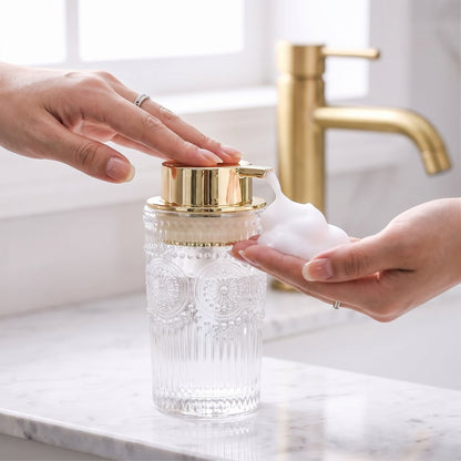 Hands pumping white foam soap from a clear glass dispenser with a gold pump on a white marble counter
