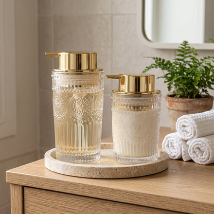 Two clear glass soap dispensers with gold pumps on a round stone tray beside rolled towels and a potted fern