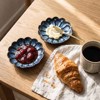 Two blue glazed ceramic dishes with dark scalloped rims, one holding strawberry jam and one holding butter with a small knife, on an oak table beside a croissant and a mug of coffee