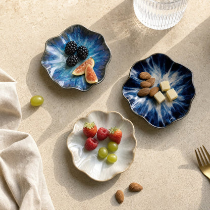 Overhead view of three glazed ceramic dishes in blue, navy and cream holding blackberries and figs, almonds and cheese cubes, and strawberries, raspberries and grapes on a beige stone surface