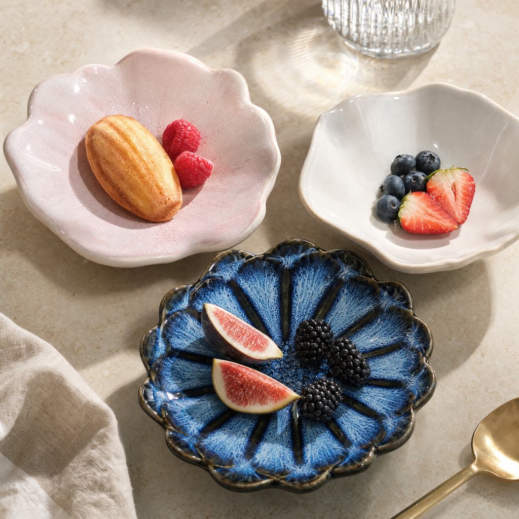 Three glazed ceramic dishes in blush pink, ivory and cobalt blue holding a madeleine and raspberries, blueberries and strawberries, and figs and blackberries on a beige stone surface