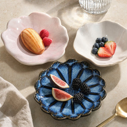 Three glazed ceramic dishes in blush pink, ivory and cobalt blue holding a madeleine and raspberries, blueberries and strawberries, and figs and blackberries on a beige stone surface