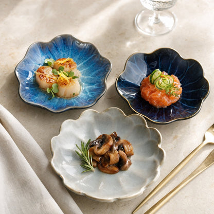 Three glazed ceramic dishes in cobalt blue, navy and pale gray-blue holding seared scallops, salmon tartare and sauteed mushrooms with a rosemary sprig on a beige surface
