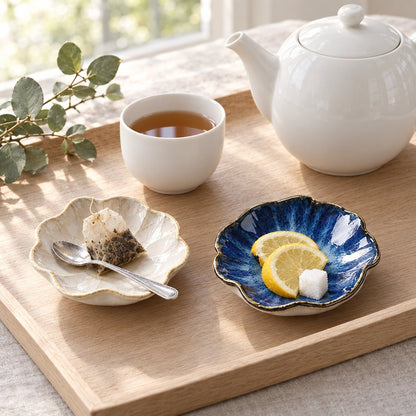 Two glazed ceramic dishes on a wooden tray, a cream crackle-glaze dish holding a used tea bag and spoon and a cobalt blue dish holding lemon slices and a sugar cube, beside a white teapot and teacup