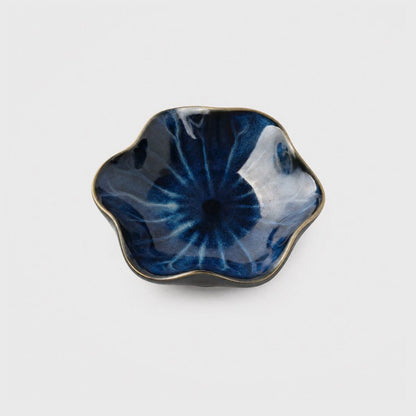 Single glossy deep navy glazed ceramic dish with pale blue streaks and a thin bronze rim in a wavy flower shape on a plain light gray background