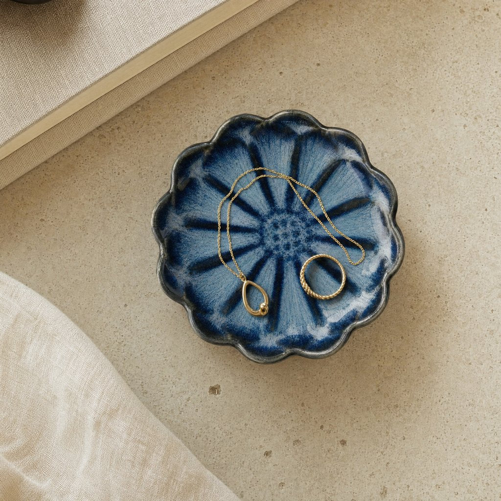 Overhead view of a blue glazed ceramic dish with radiating petal stripes and a scalloped dark rim holding a gold chain necklace and a gold ring on a beige stone surface
