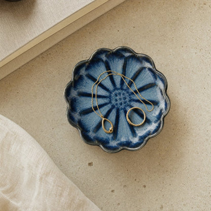 Overhead view of a blue glazed ceramic dish with radiating petal stripes and a scalloped dark rim holding a gold chain necklace and a gold ring on a beige stone surface