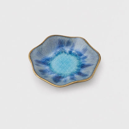 Single gray-blue glazed ceramic dish with a bright turquoise center, dark blue streaks and a thin gold rim in a wavy shape on a plain light gray background