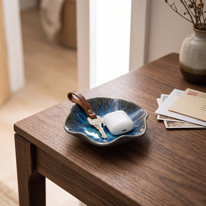 Blue and teal glazed ceramic dish with a wavy rim holding a silver key on a leather loop and a white earbud case on a walnut side table