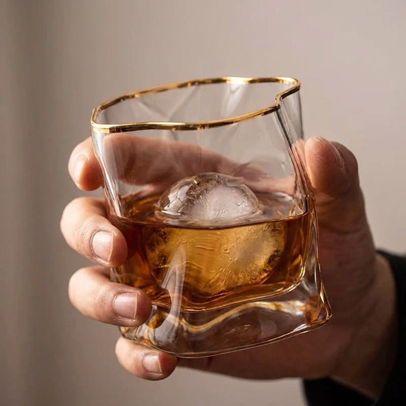 Hand holding a glass of whiskey with a gold rim against a neutral background