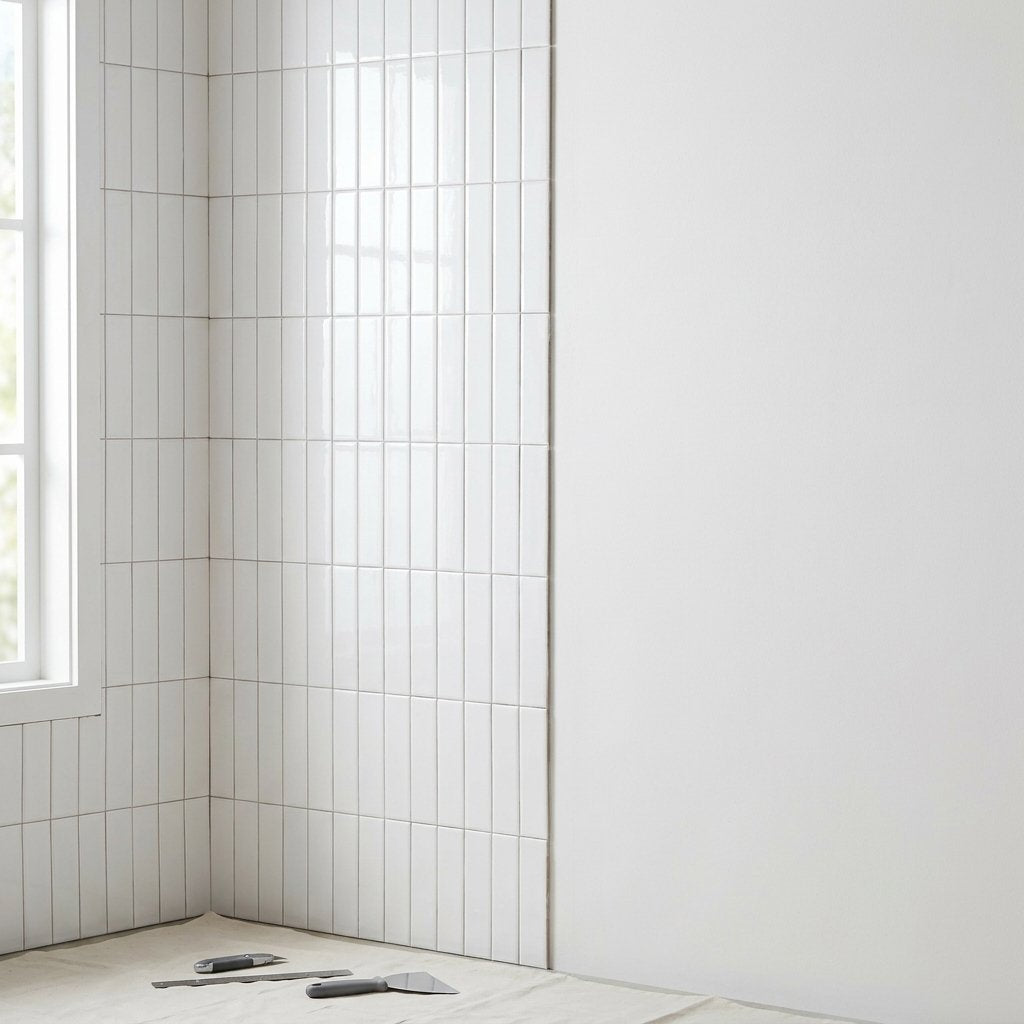 White glossy subway tile partially installed on a wall corner with a utility knife and trowel on a drop cloth