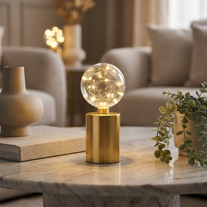 Lit clear glass globe LED lamp with gold wire fairy lights on a brass base, on a marble coffee table with books and a potted plant