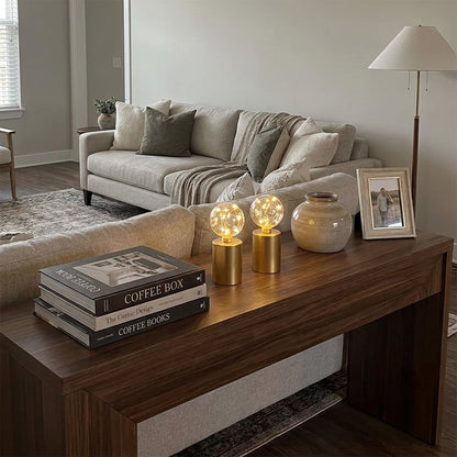 Two lit glass globe LED lamps with gold wire fairy lights on a dark wood console table with books and a vase