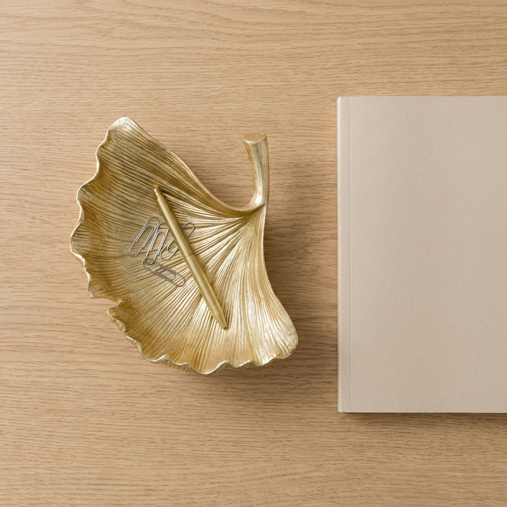 Overhead view of a gold ginkgo leaf tray holding paperclips and a pen on a desk