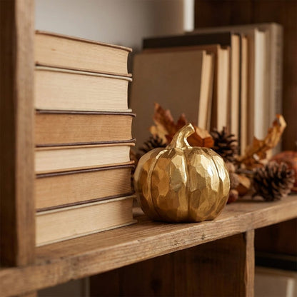 Gold faceted pumpkin on a wood bookshelf beside a stack of books, pinecones, and faux fall leaves