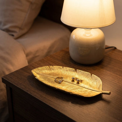 Gold leaf tray with a ring and earrings on a nightstand beside a lit lamp