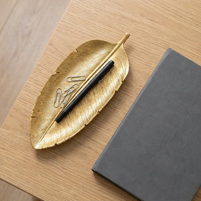 Gold leaf tray holding a pen and paperclips on a wood desk beside a notebook