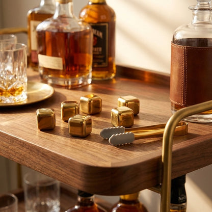 Six gold chilling stones and gold tongs on a walnut bar cart tray beside a decanter and whiskey bottles