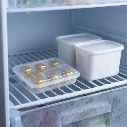 Gold chilling stones in a clear lidded tray with gold tongs on a frosty freezer shelf