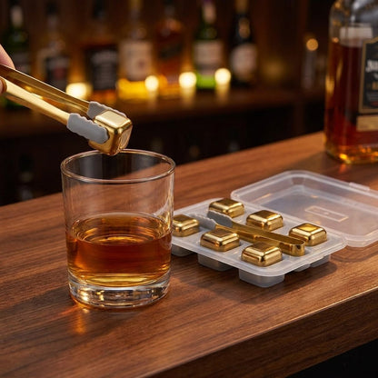 Gold chilling stones in a clear tray with tongs holding one stone above a whiskey glass at a wooden bar
