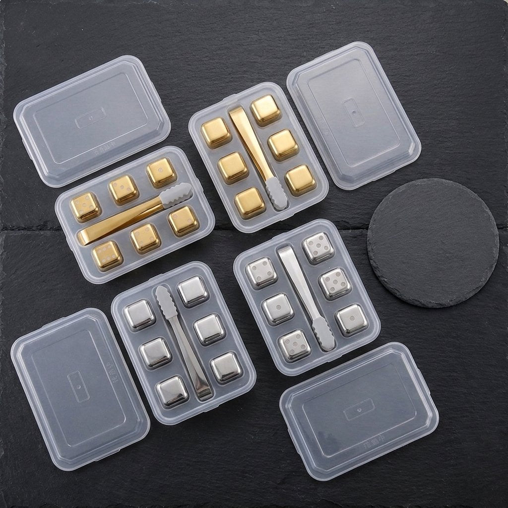 Four clear lidded trays of gold and silver chilling stones, plain and dice-style, with tongs on dark slate