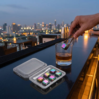 Iridescent rainbow stainless steel chilling stones in a clear tray, with one held in tongs above a whiskey tumbler at a rooftop bar
