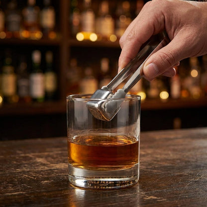 Hand holding silver tongs with a silver chilling stone above a whiskey tumbler on a wooden bar