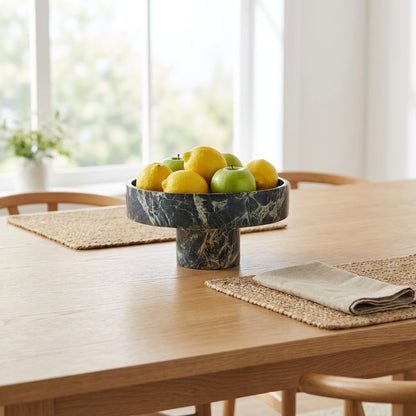 Green marble pedestal fruit bowl filled with lemons and apples on a wood dining table with woven placemats