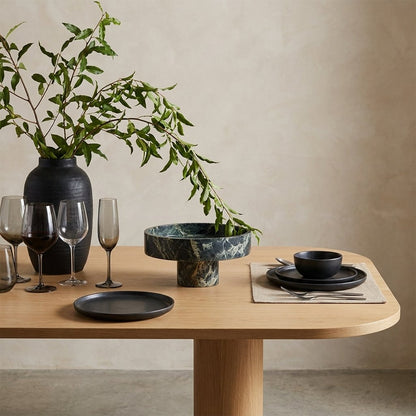 Empty green marble pedestal fruit bowl on a wood dining table beside a vase of greenery and wine glasses