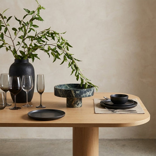 Empty green marble pedestal fruit bowl on a wood dining table beside a vase of greenery and wine glasses