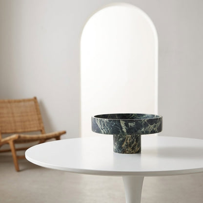 Empty green marble pedestal fruit bowl on a white round pedestal table beside a rattan chair