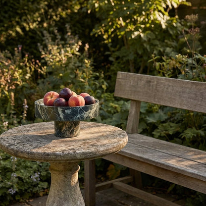 Green marble pedestal fruit bowl filled with peaches and plums on an outdoor stone pedestal table in a garden