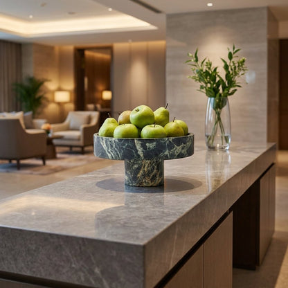 Green marble pedestal fruit bowl filled with apples and pears on a stone counter in a hotel lobby setting
