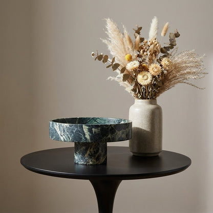 Empty green marble pedestal fruit bowl on a round side table beside a vase of dried flowers