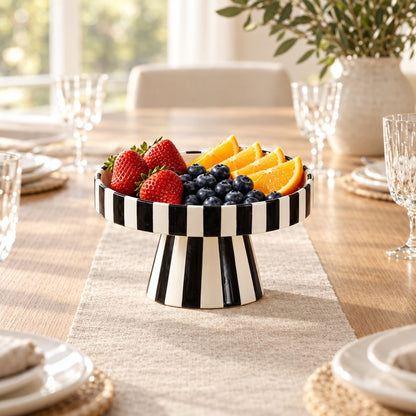 Black and white striped pedestal tray holding strawberries, blueberries and orange slices on a linen runner on a dining table with plates and glasses