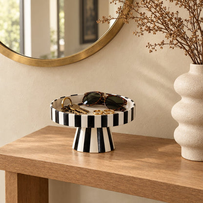 Black and white striped pedestal tray holding sunglasses and keys on a wood console table, beside a cream rippled vase with dried stems and a gold round mirror
