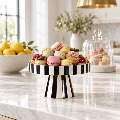 Black and white striped pedestal tray holding a pile of colorful macarons on a marble kitchen counter, with a bowl of lemons and a glass cloche in the background