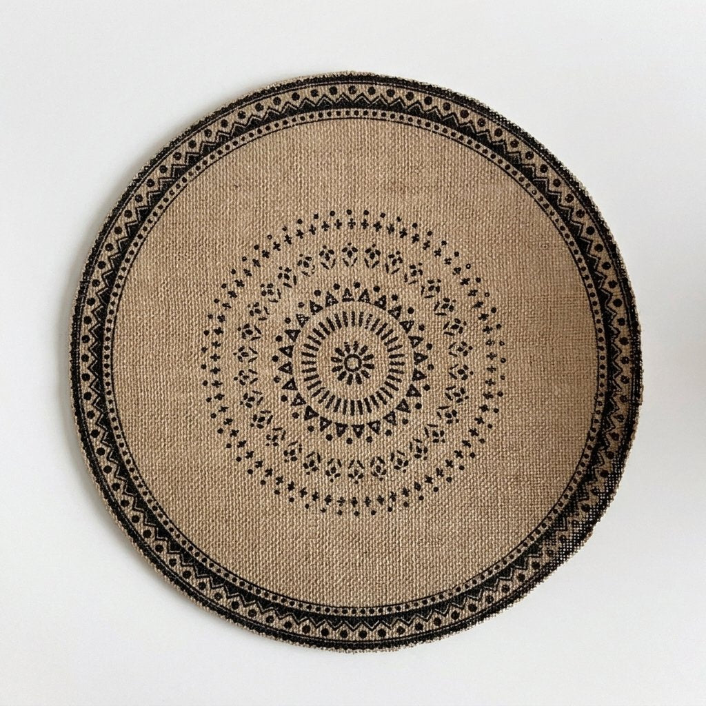 Round handwoven jute placemat with a black dotted mandala pattern and a black patterned band edge, no fringe, on a light grey background
