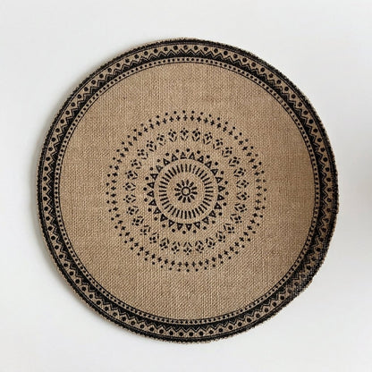 Round handwoven jute placemat with a black dotted mandala pattern and a black patterned band edge, no fringe, on a light grey background