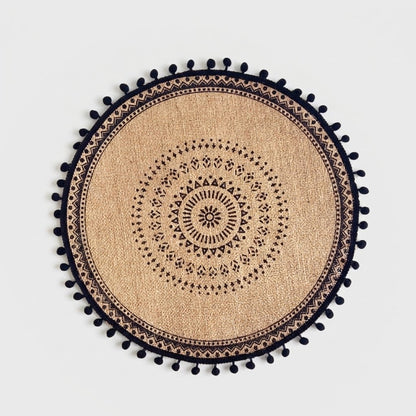 Round handwoven jute placemat with a black dotted mandala pattern and black pompom trim on a light grey background