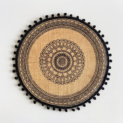 Round handwoven jute placemat with a black mandala pattern and black pompom trim on a light grey background