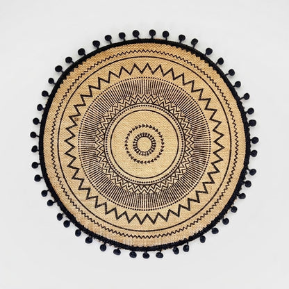 Round handwoven jute placemat with a black zigzag sunburst pattern and black pompom trim on a light grey background
