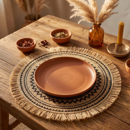 Round black zigzag pattern jute placemat with fringe under a terracotta plate on a wood table with small spice bowls, pampas grass in an amber glass vase and a beeswax candle