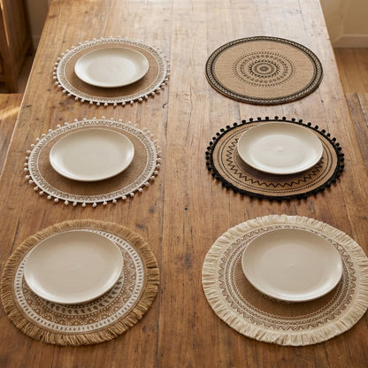 Overhead view of six round handwoven jute placemats in different designs on a wood table, five set with plates and one empty