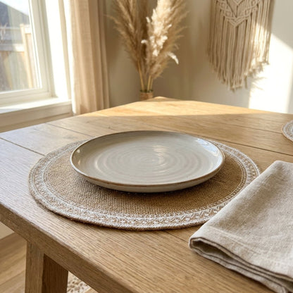 Round jute placemat with a pale lace trim edge under a speckled plate with a linen napkin on an oak table beside pampas grass and a macrame wall hanging