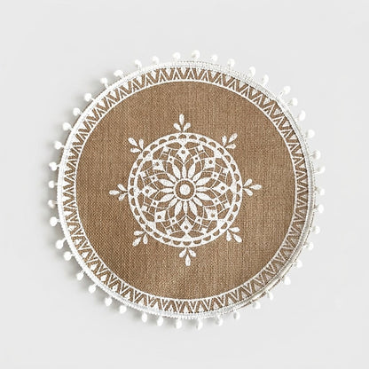 Round handwoven jute placemat with a white flower mandala pattern, zigzag band and white pompom trim on a light grey background