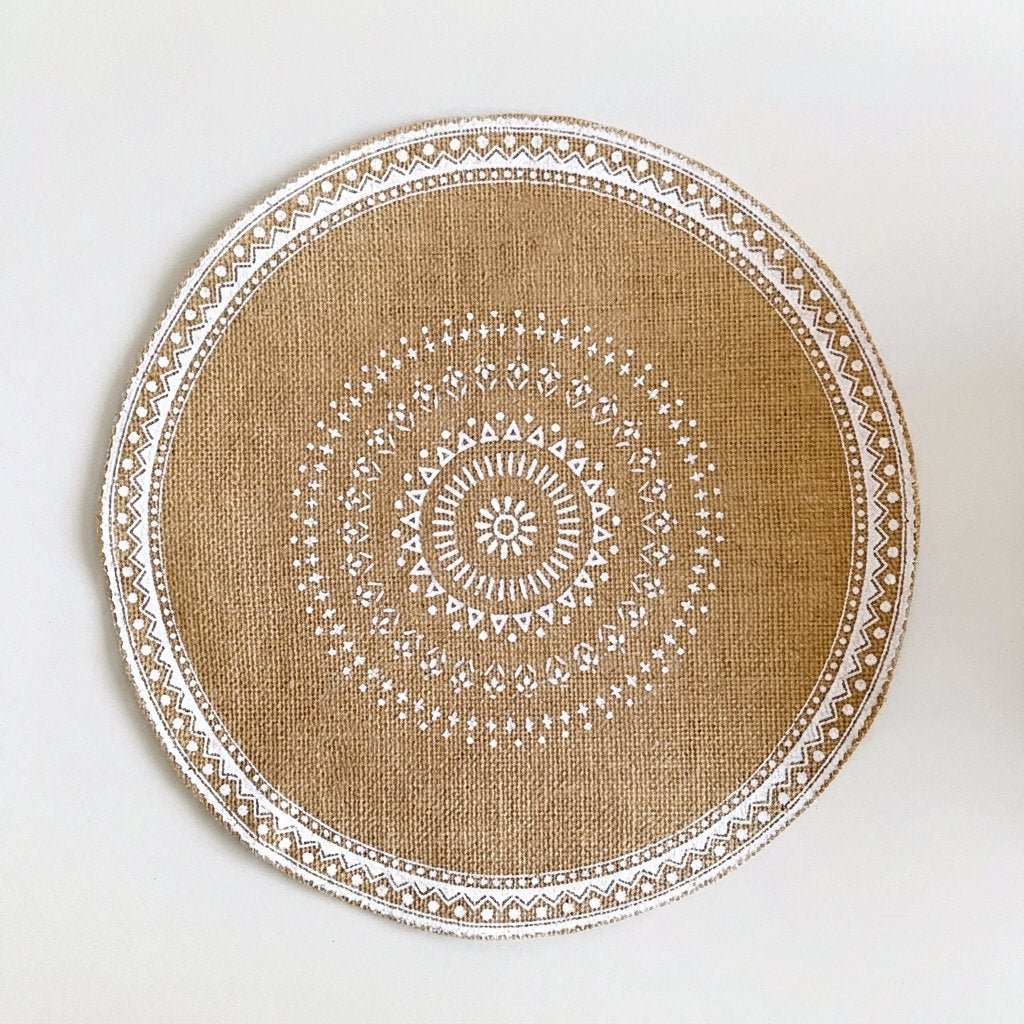 Round handwoven jute placemat with a white dotted mandala pattern and white trim edge, no fringe, on a light grey background