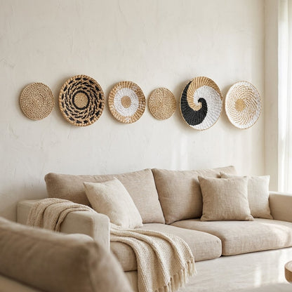 Six round woven wall baskets in tan, black and white patterns hung in a row on a cream plaster wall above a beige sofa with linen cushions and a chunky knit throw