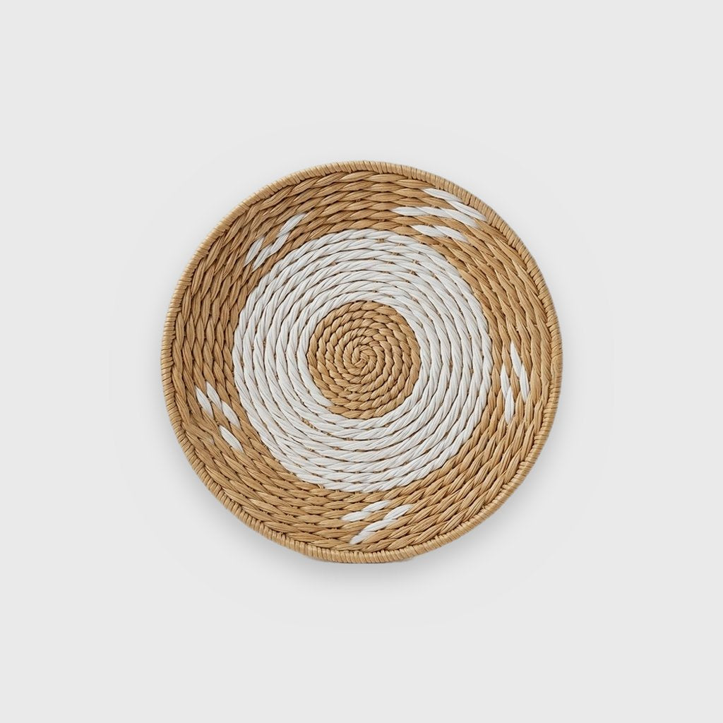 Single shallow round woven basket with a tan center spiral, a wide white ring and small white dashes on a tan outer band, on a plain light gray background