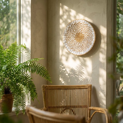 Round white and tan woven wall basket hung high on a sunlit beige wall above a potted fern on a windowsill